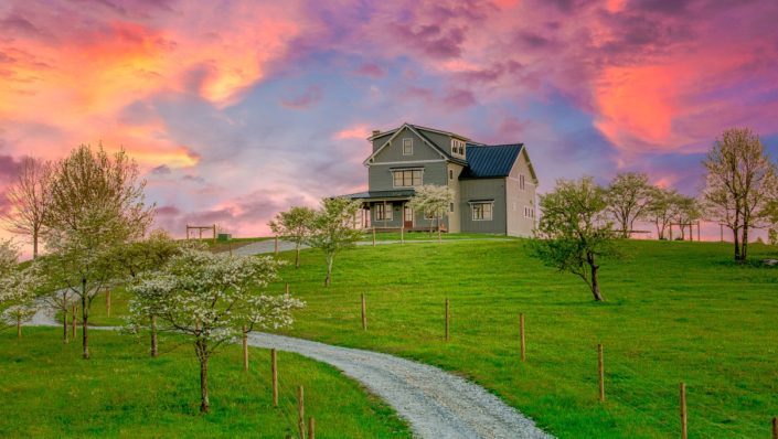 The exterior of a modern home with a wrap-around porch at sunset. The colors of the sunset are vibrant pink, purple, orange, and yellow. There is a gravel driveway that leads up to the house.